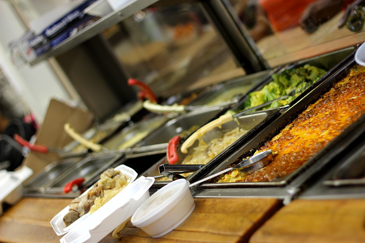 Baked mac and cheese with a browned crust, broccoli and cabbage in steam table pans, with takeout containers filled and waiting